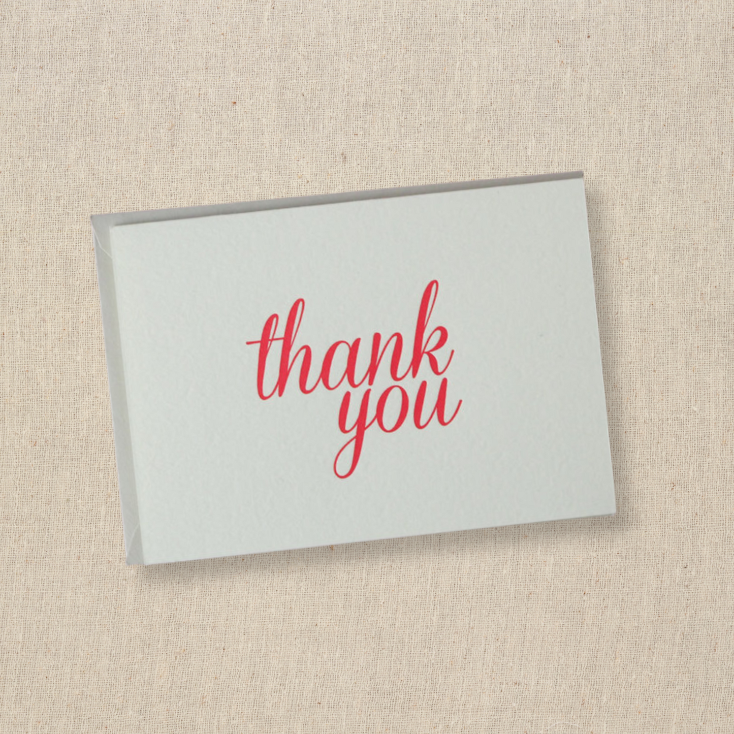 Notecard in cotton paper with the words thank you done in modern script in red ink Light gray envelope is underneath card.