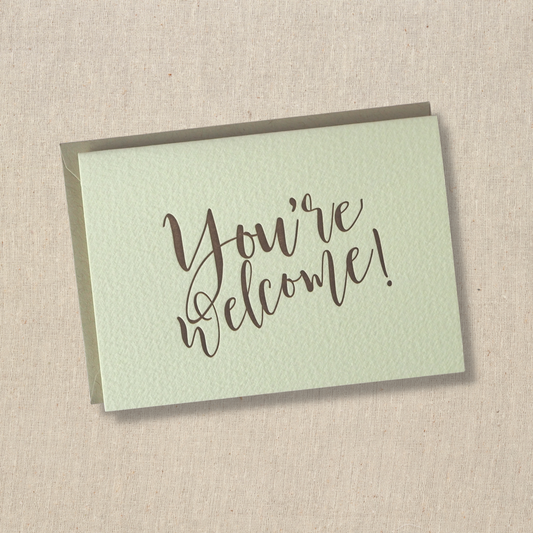 Notecard with the words you're welcome printed in brown ink with modern cursive script. Light kraft envelope underneath