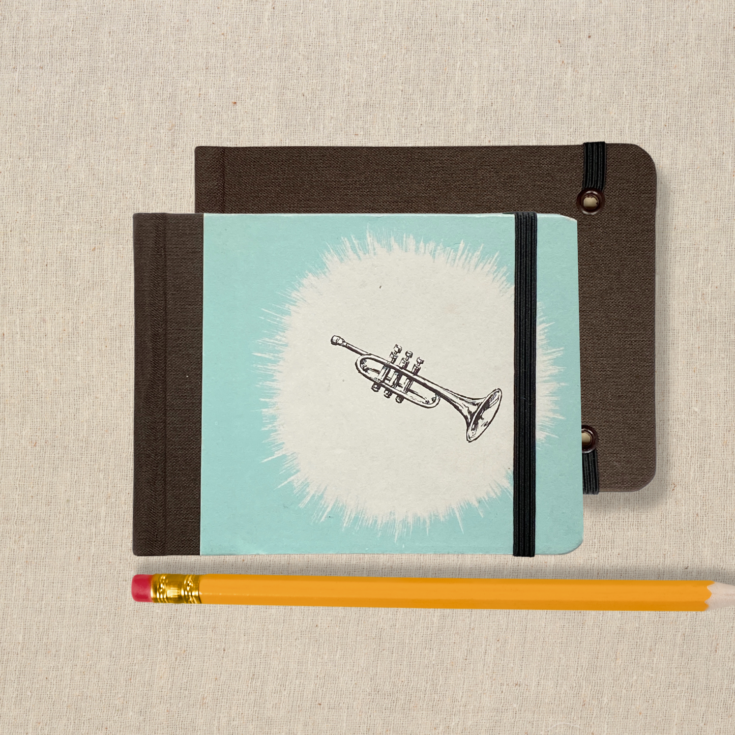 Small hand bound sketchbook with vintage illustration of black and trumpet against a white starburst on a light blue background on front cover. Spine and back cover are dark brown book cloth. Book closes with black elastic.