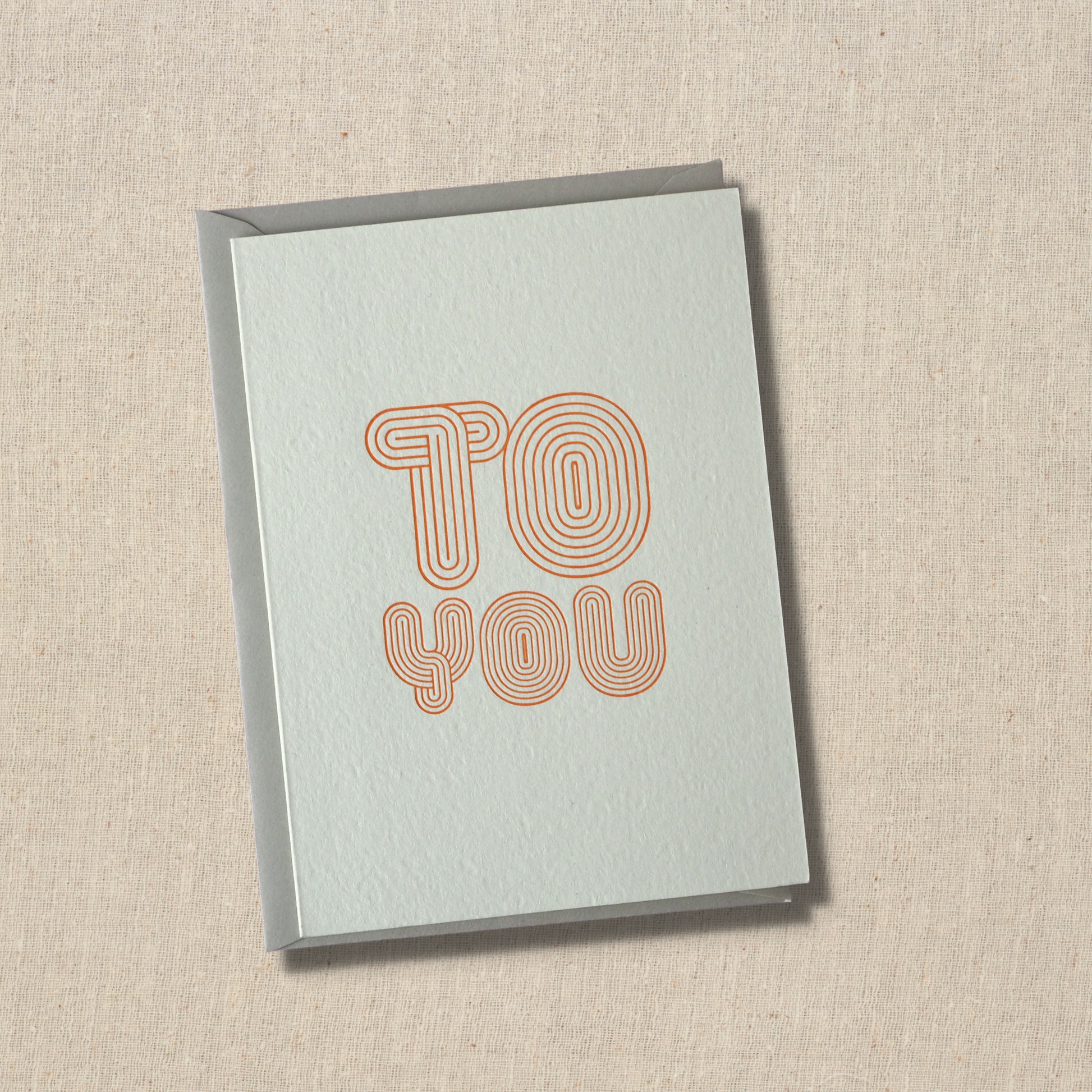 Notecard with the words to you printed on front. Type face  looks like bubble letters with a series of lines inside. Like a maze. Light gray envelope underneath.