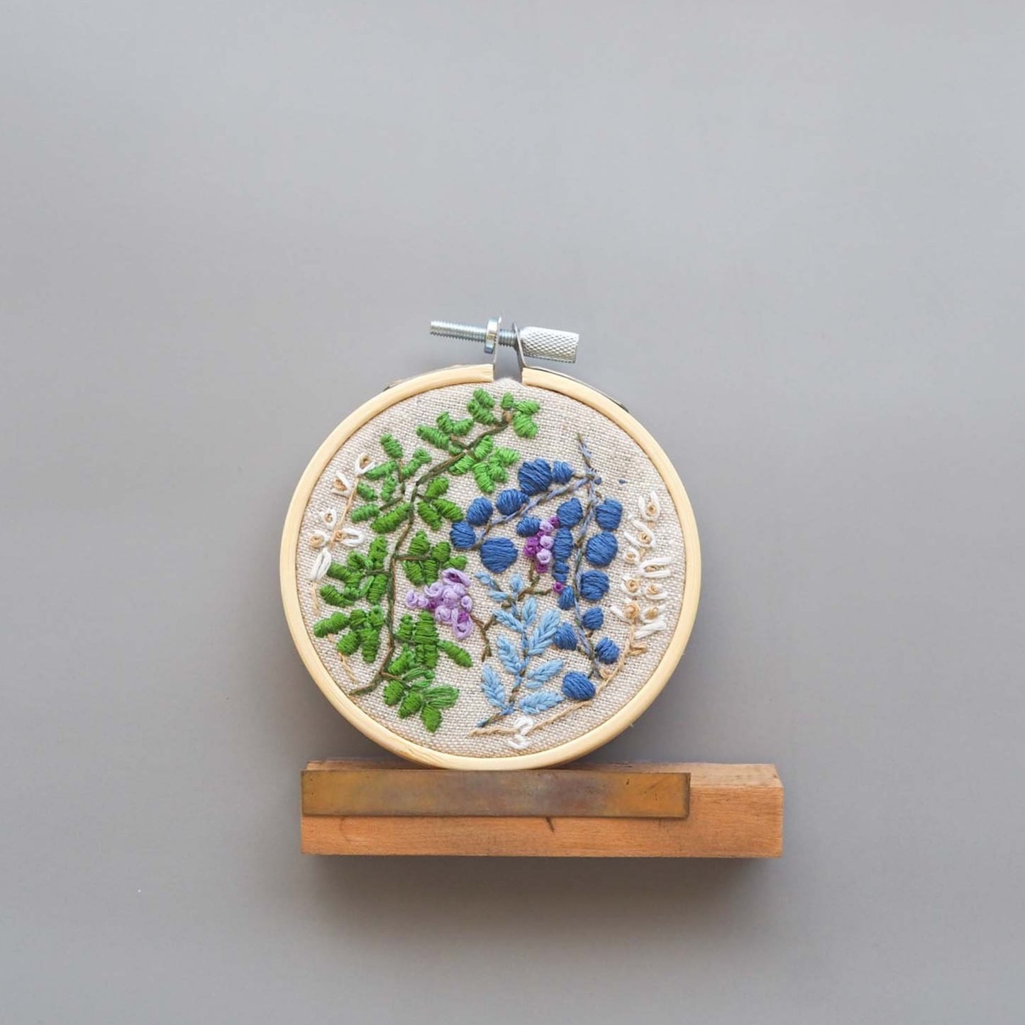 Various small leaves and floral buds in green, blue, and purple stitched on natural linen stretched on a 3 in hoop