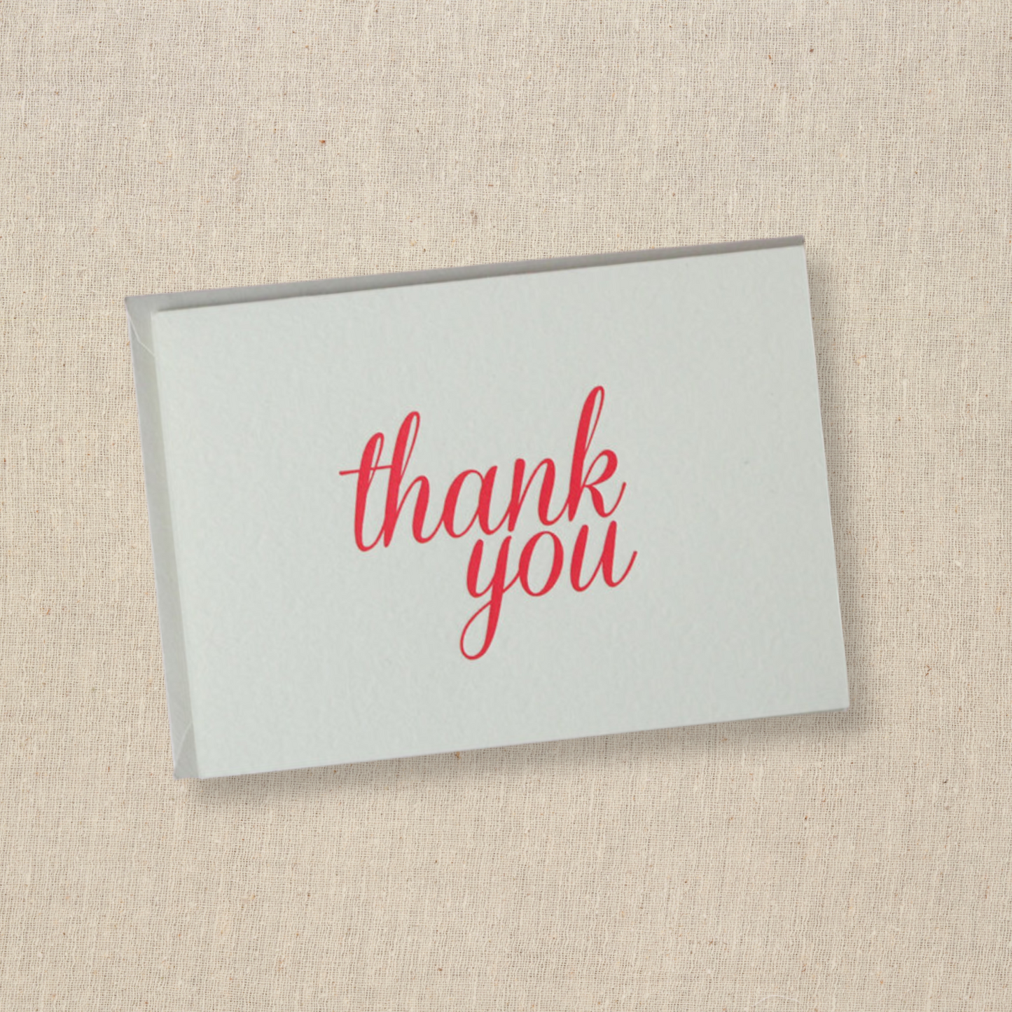 Notecard in cotton paper with the words thank you done in modern script in red ink  Light gray envelope is underneath card.