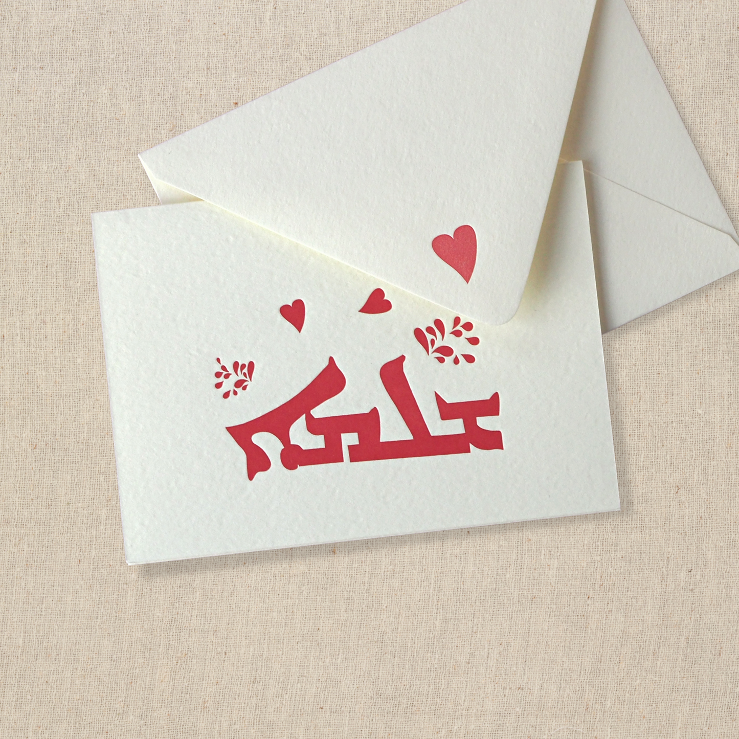 Notecard on cotton paper. Red text in syriac with red hearts. Transliteration is shlama. Translates to hello in English. White envelope with red heart printed on back flap is next to card.