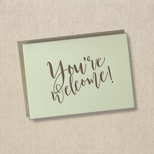 Notecard with the words you're welcome printed in brown ink with modern cursive script. Light kraft envelope underneath 